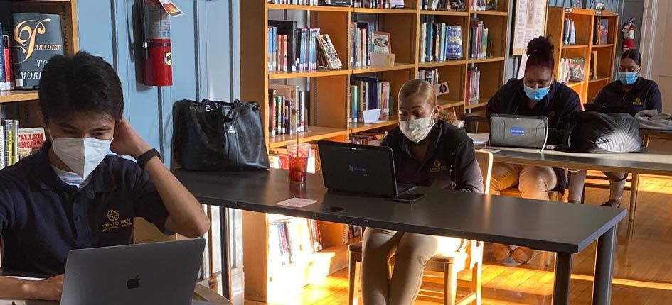 四个学生，一男三女，在图书馆学习。每个人都在自己的办公桌前，相隔6英尺，戴着口罩，用笔记本电脑工作。
