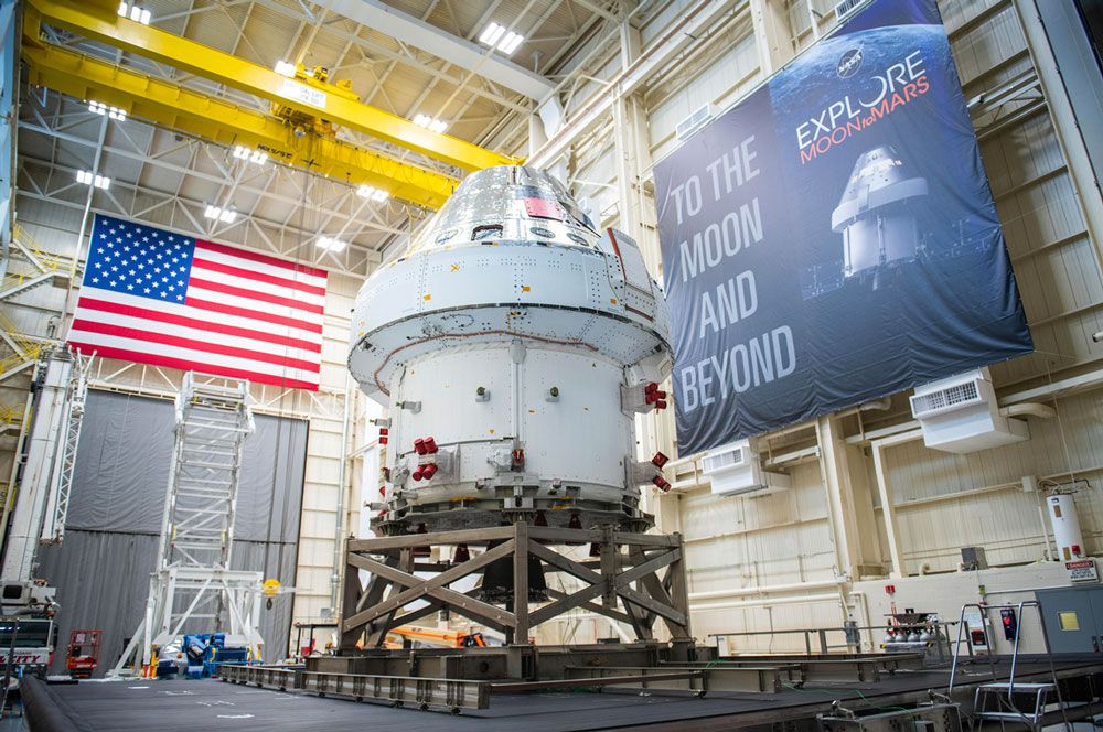 猎户座飞船在一个大型设施内，一面墙上挂着美国国旗，另一面墙上挂着横幅，上面写着“奔向月球和更远的地方”。